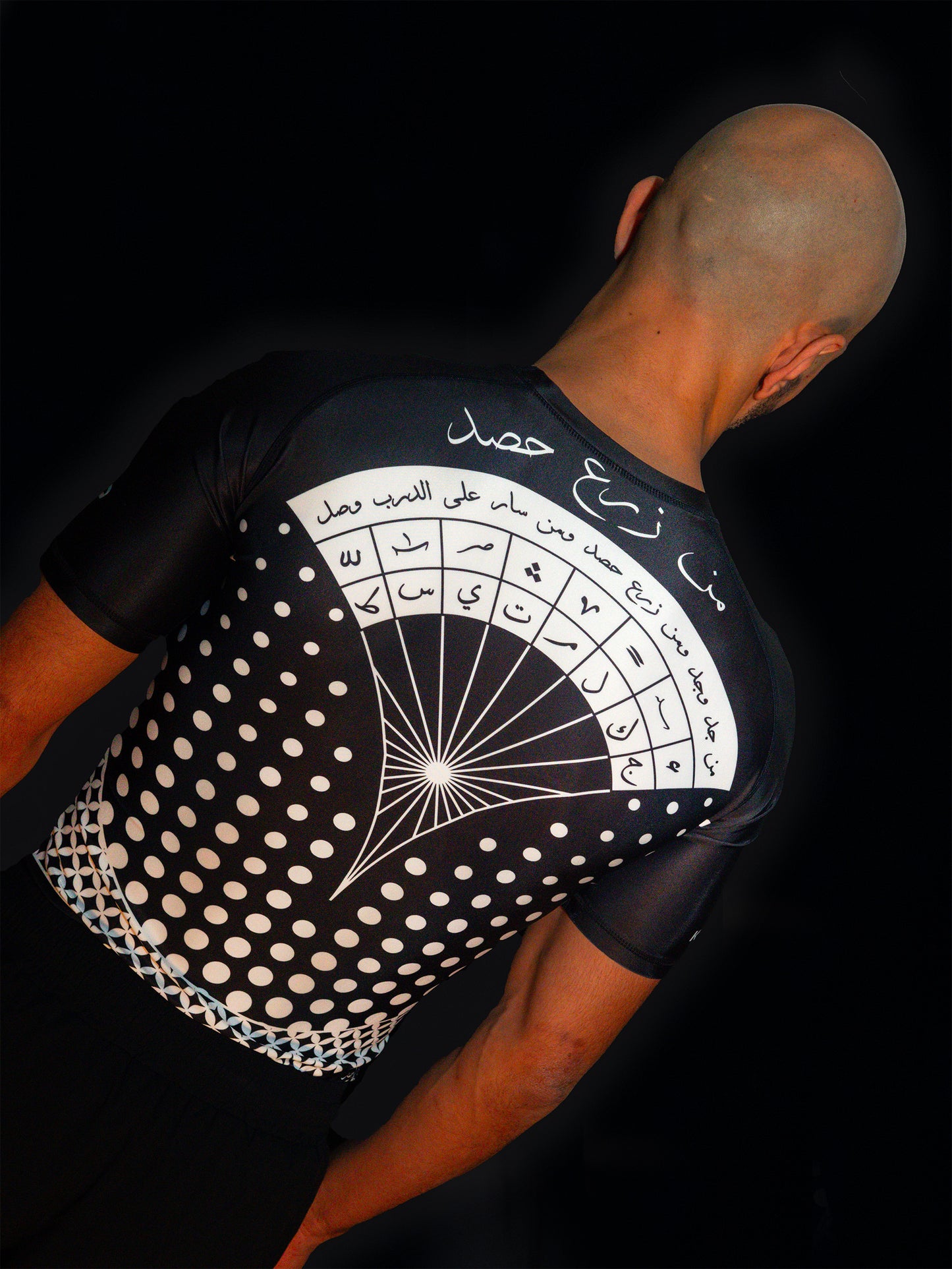 Arab model wearing black rashguard with arabic design, used for jiu-jitsu and other martial arts (e.g. MMA). This product features an exclusive print about a popular, common arabic expression regarding work, perseverance and achievement. Using thuluth, a type of arabic calligraphy, it exhibits a beautiful, geometrical style, featuring arabic lettering and symbols. It also gives a sensation of interstellar and space travel. Back view, black and white print color. Backside view.