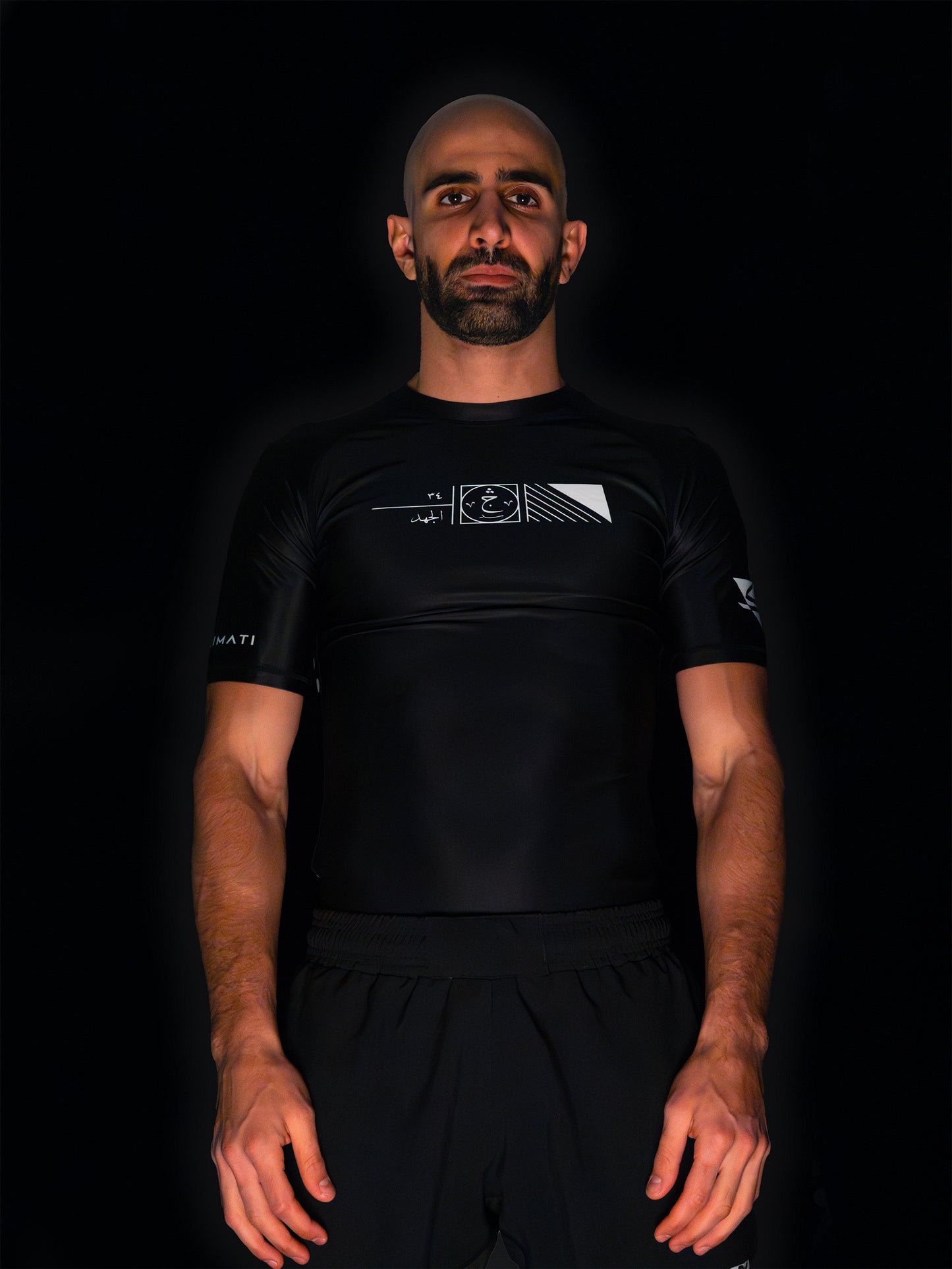 Arab model wearing black rashguard with arabic design, used for jiu-jitsu and other martial arts (e.g. MMA). This product features an exclusive print about a popular, common arabic expression regarding work, perseverance and achievement. Using thuluth, a type of arabic calligraphy, it exhibits a beautiful, geometrical style, featuring arabic lettering and symbols. It also gives a sensation of interstellar and space travel. Back view, black and white print color. Front view.
