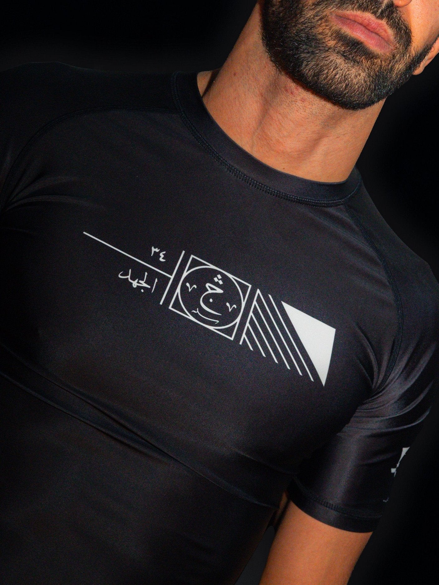 Arab model wearing black rashguard with arabic design, used for jiu-jitsu and other martial arts (e.g. MMA). This product features an exclusive print about a popular, common arabic expression regarding work, perseverance and achievement. Using thuluth, a type of arabic calligraphy, it exhibits a beautiful, geometrical style, featuring arabic lettering and symbols. It also gives a sensation of interstellar and space travel. Back view, black and white print color. Frontside close-up view.