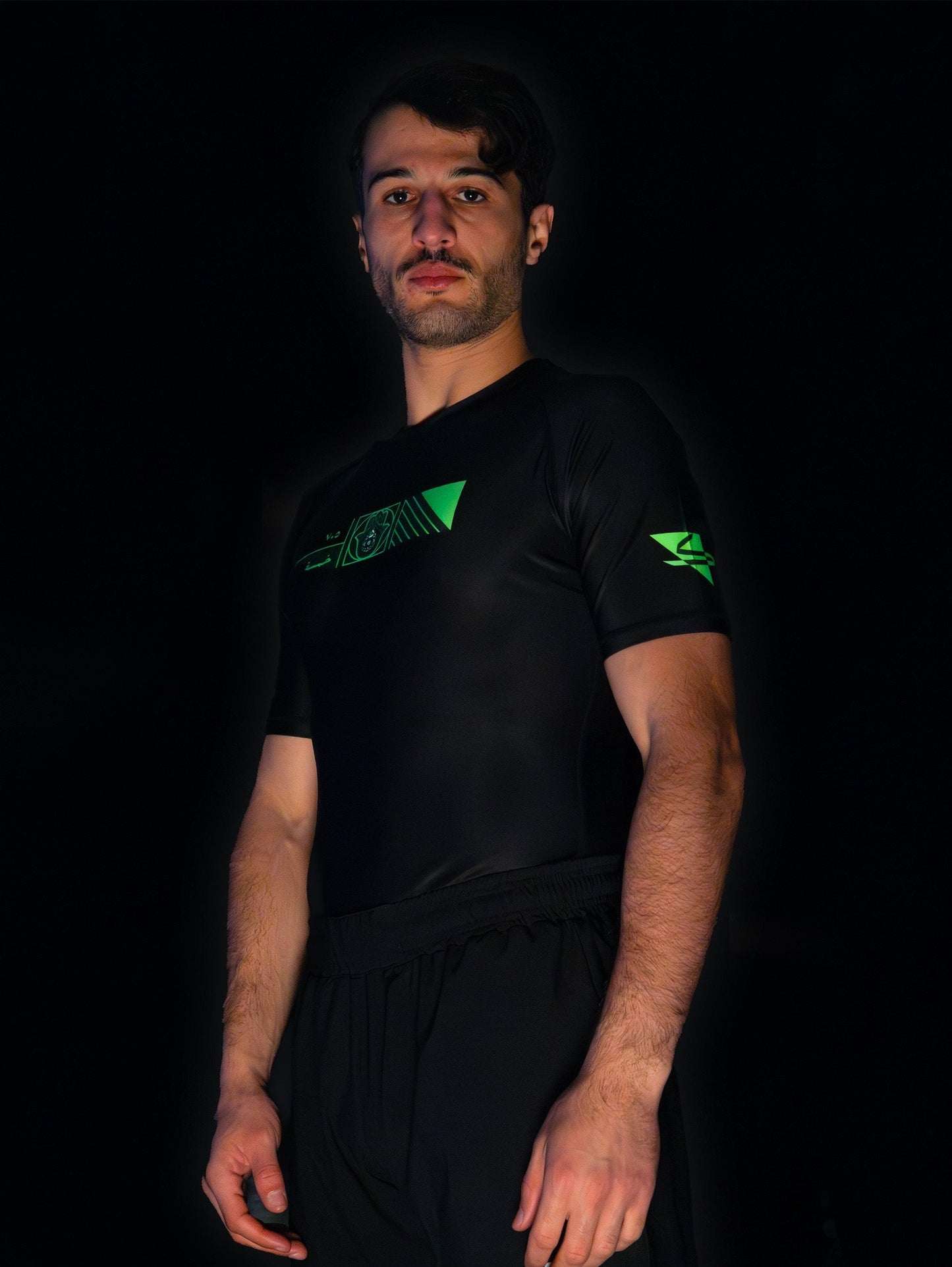 Black rashguard with arabic design, used for jiu-jitsu and other martial arts (e.g. MMA). This product features an exclusive print about khamsa, also known as hamsa, or the hand of Fatima. It is a popular amulet in Middle Eastern and North African culture, and is intended to protect against evil and envy. This art piece and pattern uses beautiful arabic letters and calligraphy to form its attractive shape. Yellow and white print color. Arab model wearing it. left side wide picture.