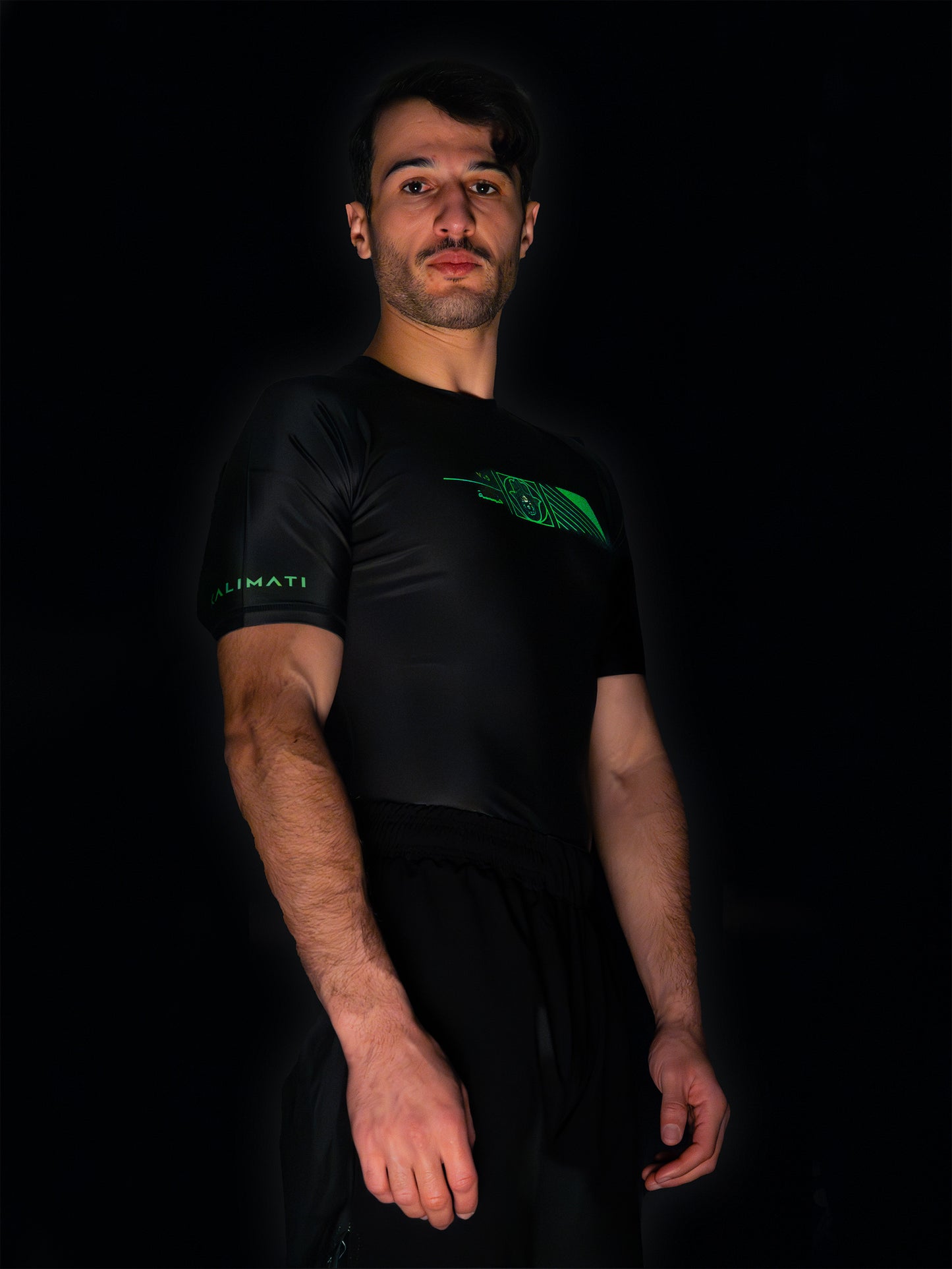 Black rashguard with arabic design, used for jiu-jitsu and other martial arts (e.g. MMA). This product features an exclusive print about khamsa, also known as hamsa, or the hand of Fatima. It is a popular amulet in Middle Eastern and North African culture, and is intended to protect against evil and envy. This art piece and pattern uses beautiful arabic letters and calligraphy to form its attractive shape. Yellow and white print color. Arab model wearing it. Right side wide picture.