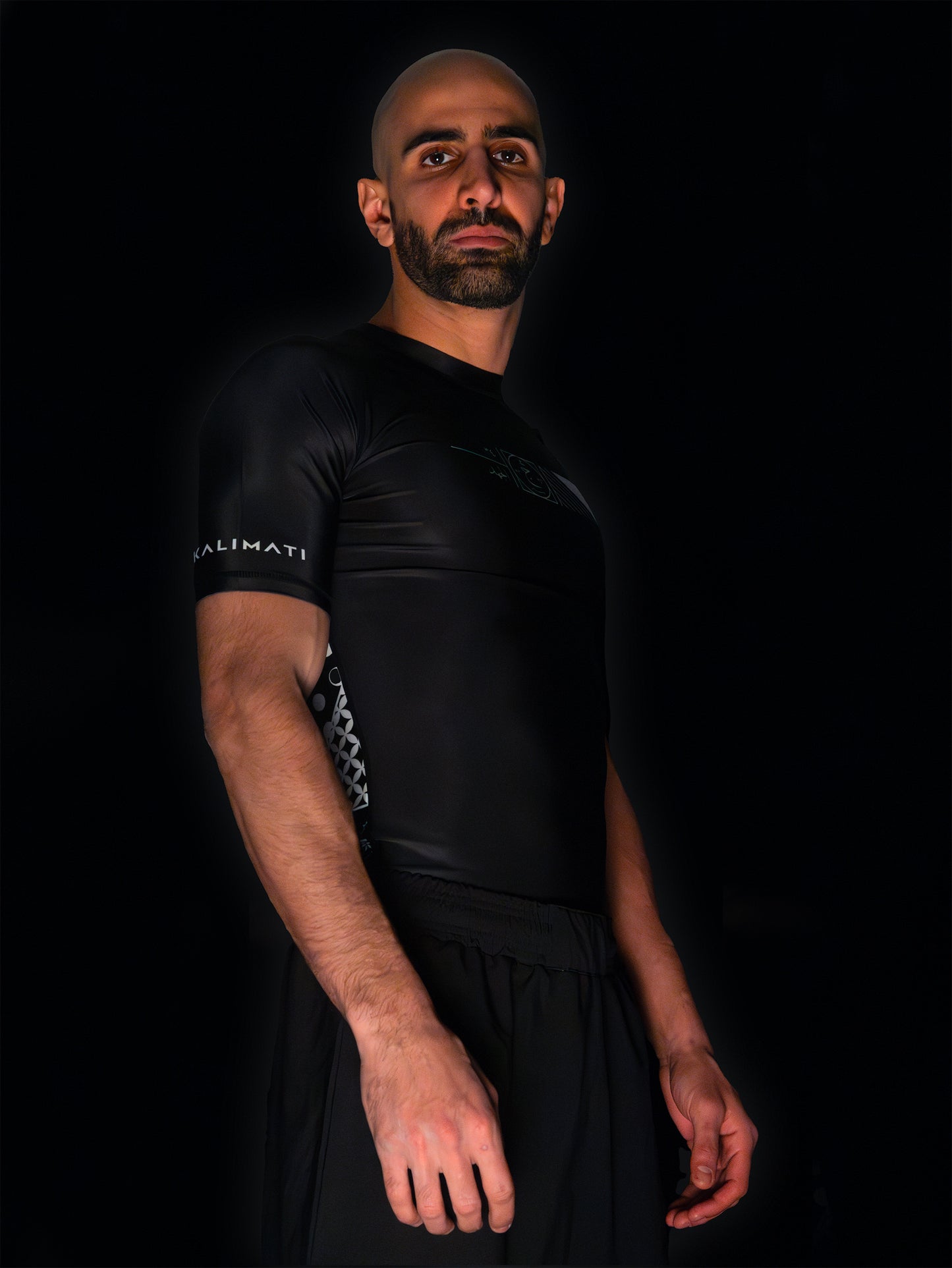 Arab model wearing black rashguard with arabic design, used for jiu-jitsu and other martial arts (e.g. MMA). This product features an exclusive print about a popular, common arabic expression regarding work, perseverance and achievement. Using thuluth, a type of arabic calligraphy, it exhibits a beautiful, geometrical style, featuring arabic lettering and symbols. It also gives a sensation of interstellar and space travel. Back view, black and white print color. Sideaways view.