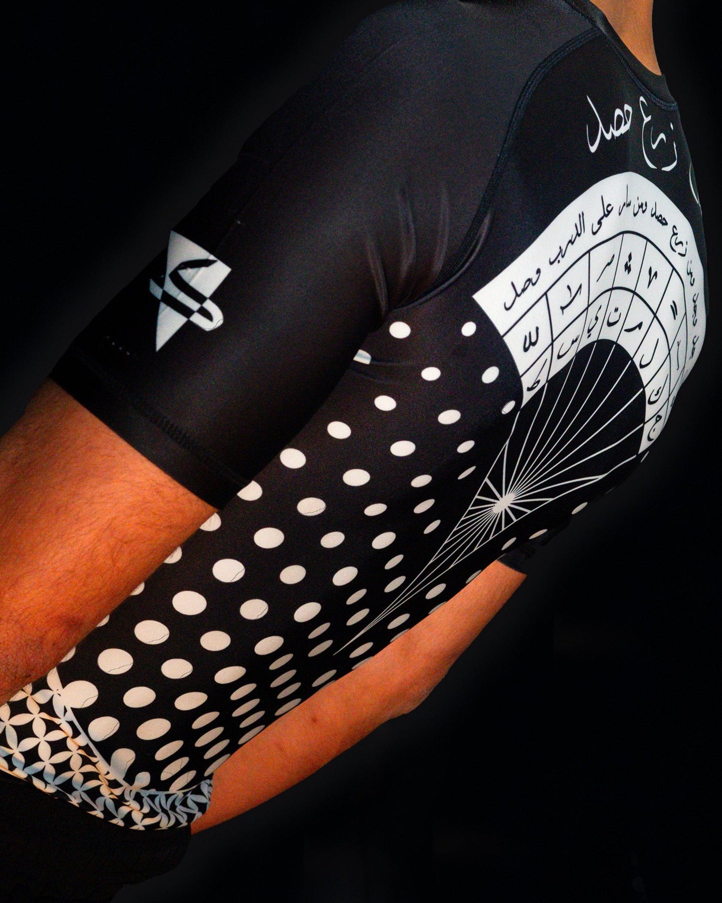 Arab model wearing black rashguard with arabic design, used for jiu-jitsu and other martial arts (e.g. MMA). This product features an exclusive print about a popular, common arabic expression regarding work, perseverance and achievement. Using thuluth, a type of arabic calligraphy, it exhibits a beautiful, geometrical style, featuring arabic lettering and symbols. It also gives a sensation of interstellar and space travel. Back view, black and white print color. Sideaways close-up view.