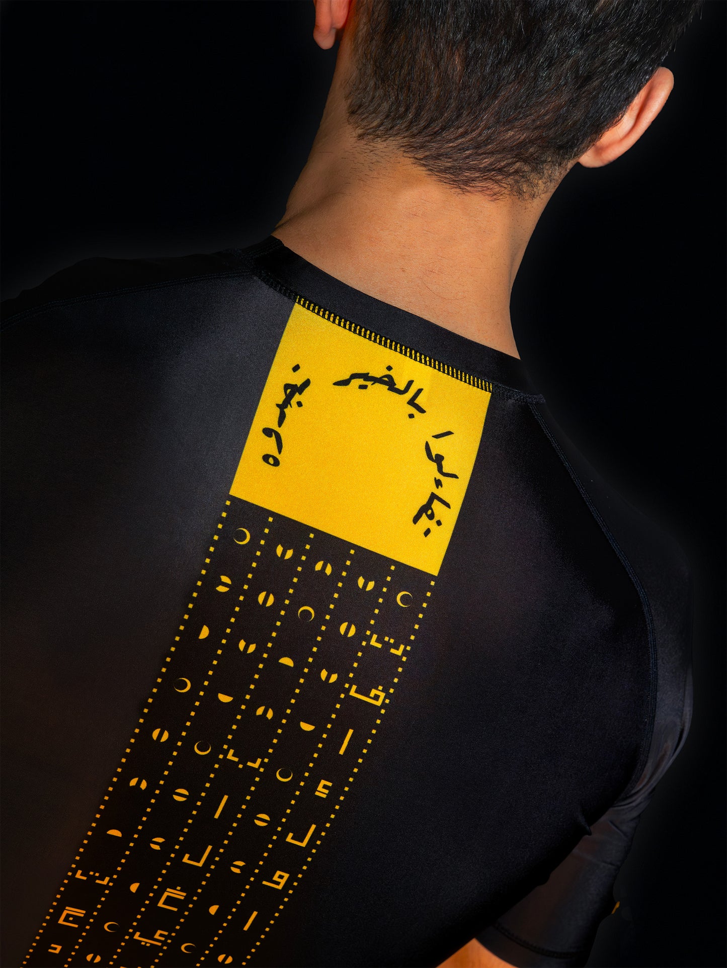 Black rashguard with arabic design, used for jiu-jitsu and other martial arts (e.g. MMA). This product features an exclusive print about a popular, common arabic expression regarding hope and optimism. Inspired by different types of arabic calligraphy, it exhibits a beautiful, geometrical style, featuring popular symbols such as the crescent, moon and stars. Backside view. Yellow and Green print color. Close-up