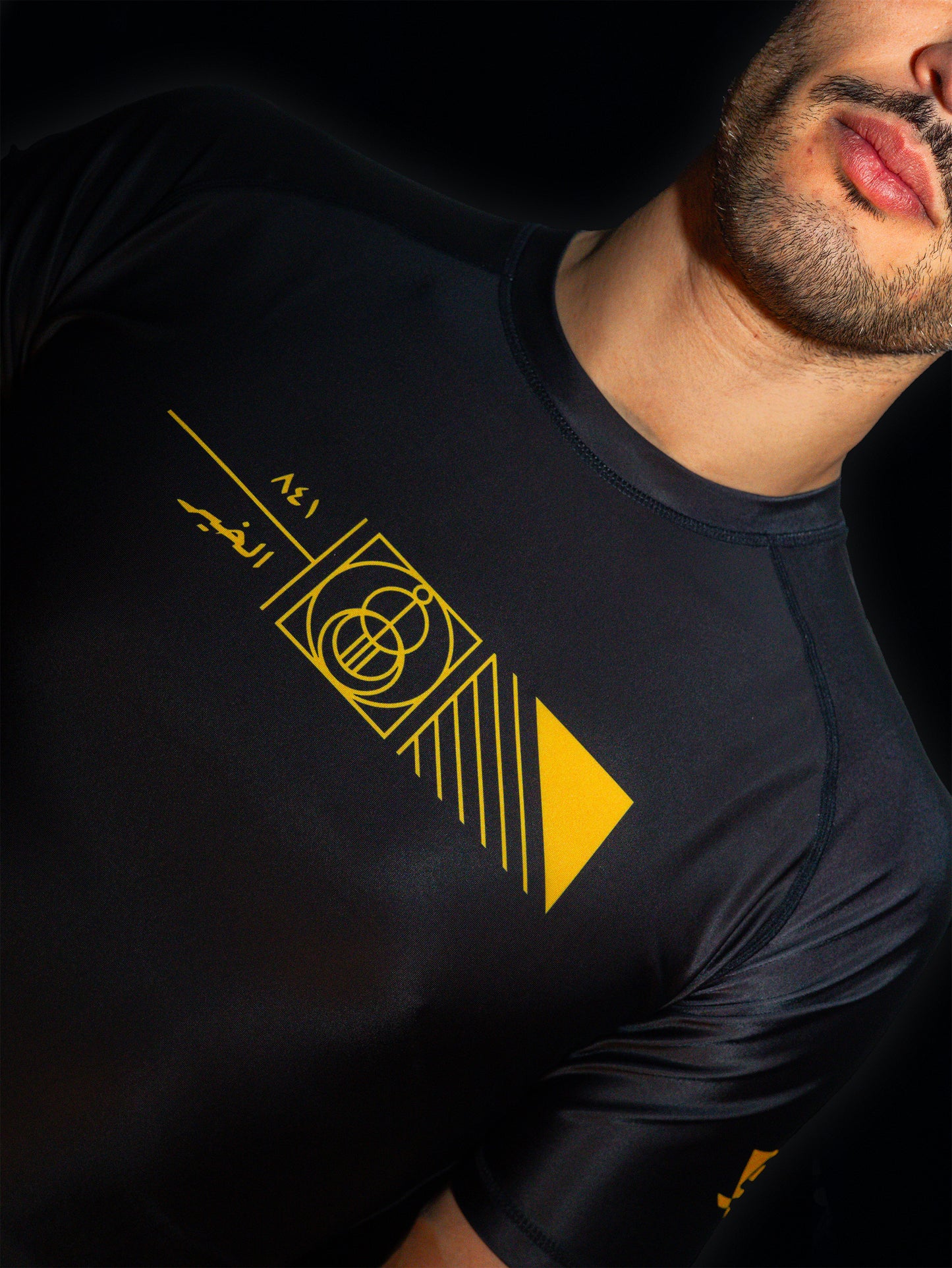 Black rashguard with arabic design, used for jiu-jitsu and other martial arts (e.g. MMA). This product features an exclusive print about a popular, common arabic expression regarding hope and optimism. Inspired by different types of arabic calligraphy, it exhibits a beautiful, geometrical style, featuring popular symbols such as the crescent, moon and stars. Frontside view. Yellow and Green print color. Close-up