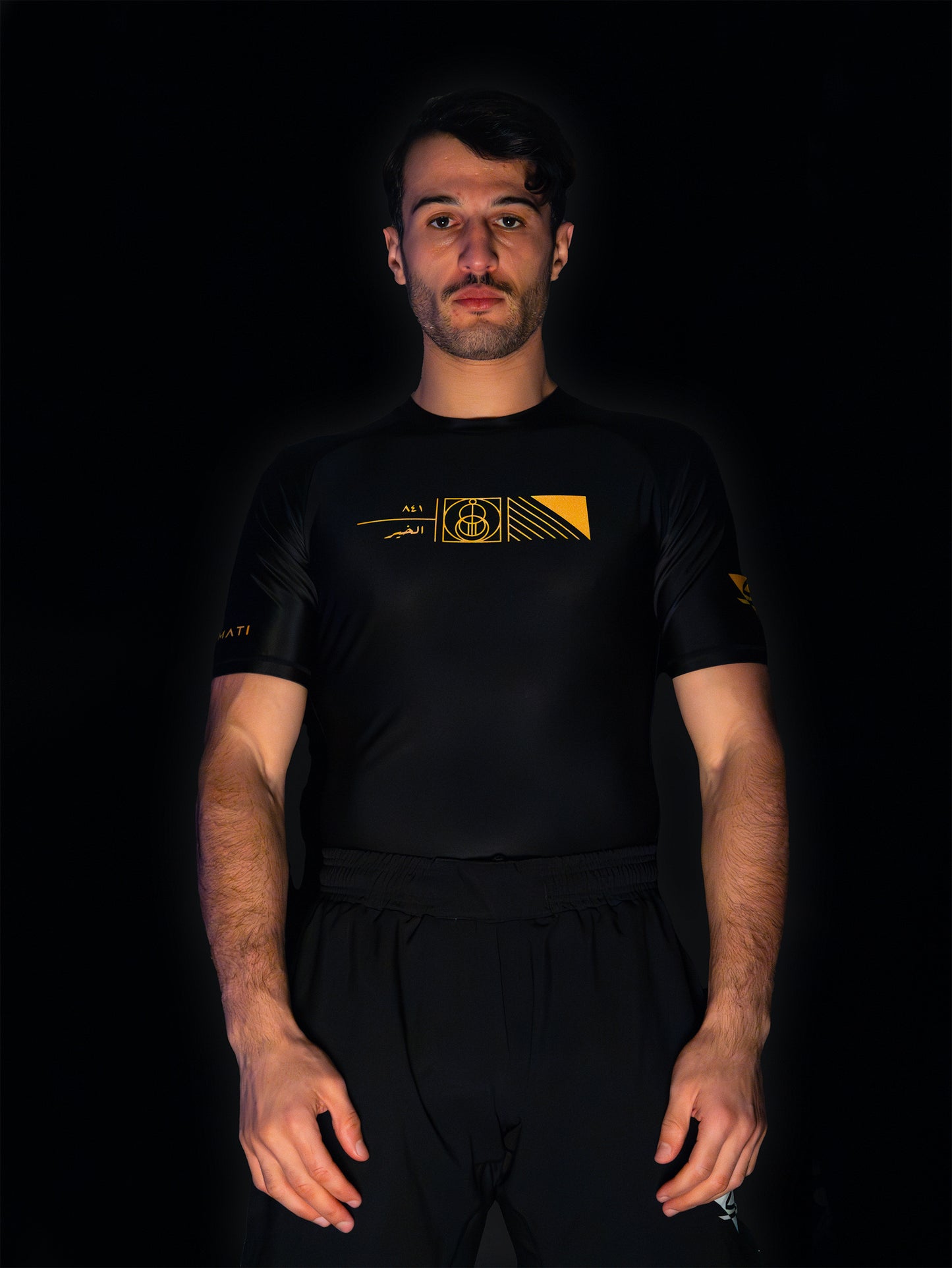 Black rashguard with arabic design, used for jiu-jitsu and other martial arts (e.g. MMA). This product features an exclusive print about a popular, common arabic expression regarding hope and optimism. Inspired by different types of arabic calligraphy, it exhibits a beautiful, geometrical style, featuring popular symbols such as the crescent, moon and stars. Frontside view. Yellow and Green print color. Wide angle.