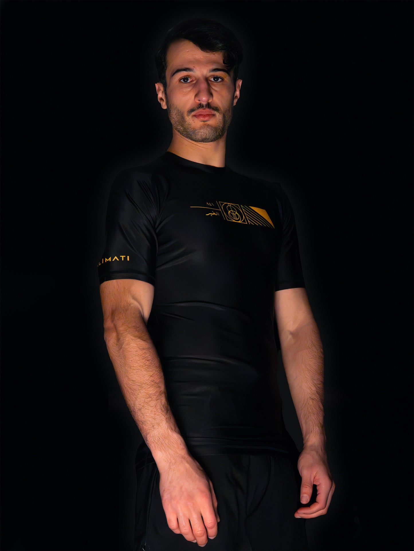 Black rashguard with arabic design, used for jiu-jitsu and other martial arts (e.g. MMA). This product features an exclusive print about a popular, common arabic expression regarding hope and optimism. Inspired by different types of arabic calligraphy, it exhibits a beautiful, geometrical style, featuring popular symbols such as the crescent, moon and stars. Frontside view. Yellow and Green print color. Right side angle.
