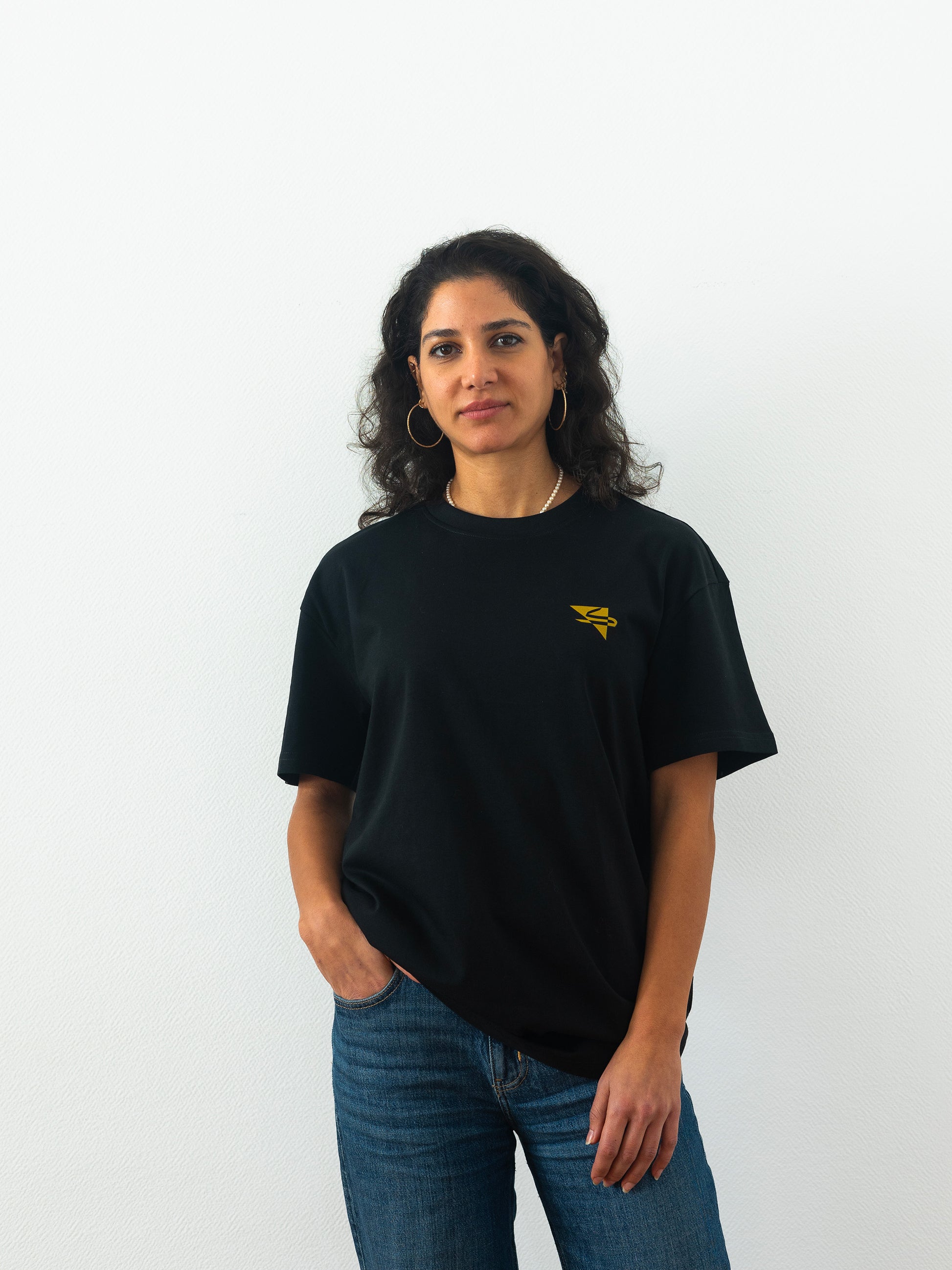 Beautiful arab model wearing a cool and creative over-sized t-shirt for streetwear and everyday lifestyle clothing. It features a simple beautiful logo, which is geometric in design and that has an arabic letter within it.  Black t-shirt. Frontside view. Black and yellow, gold print color.