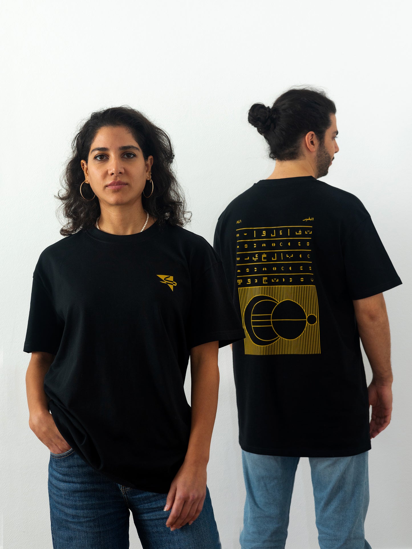 Two beautiful arab models wearing cool and creative over-sized t-shirts for streetwear and everyday lifestyle clothing. This product features an exclusive print about a popular, common arabic expression regarding hope and optimism. Inspired by different types of arabic calligraphy, it exhibits a beautiful, geometrical style, featuring popular symbols such as the crescent, moon and stars. Black t-shirt. Backside view. Black, yellow, green and gold print color. 