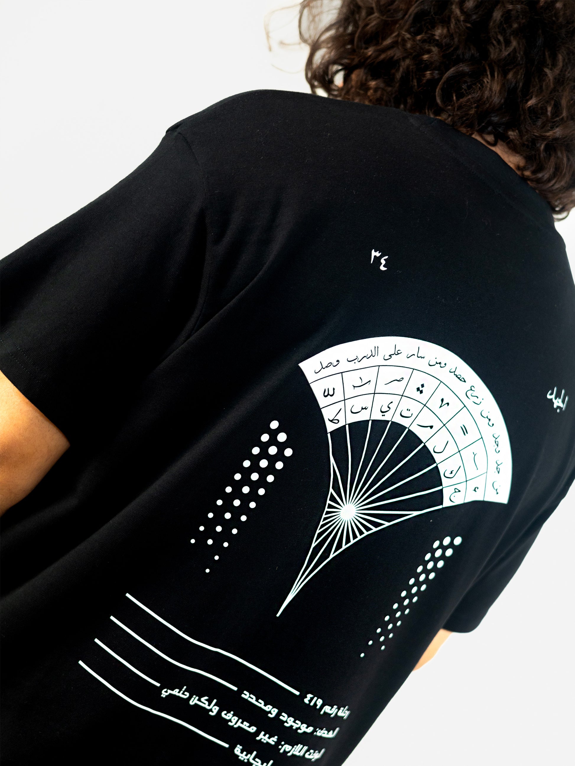 Closeup of a cool and creative over-sized t-shirt for streetwear and everyday lifestyle clothing. It features a beautiful arabic graphic print, emphasizing arabic art, culture and identity. It contains a famous arabic expression about hard work, perseverance and achievement. It uses arabic thuluth, a style of arabic calligraphy and lettering, including popular symbols. Black t-shirt. Backside view. Black and white print color.
