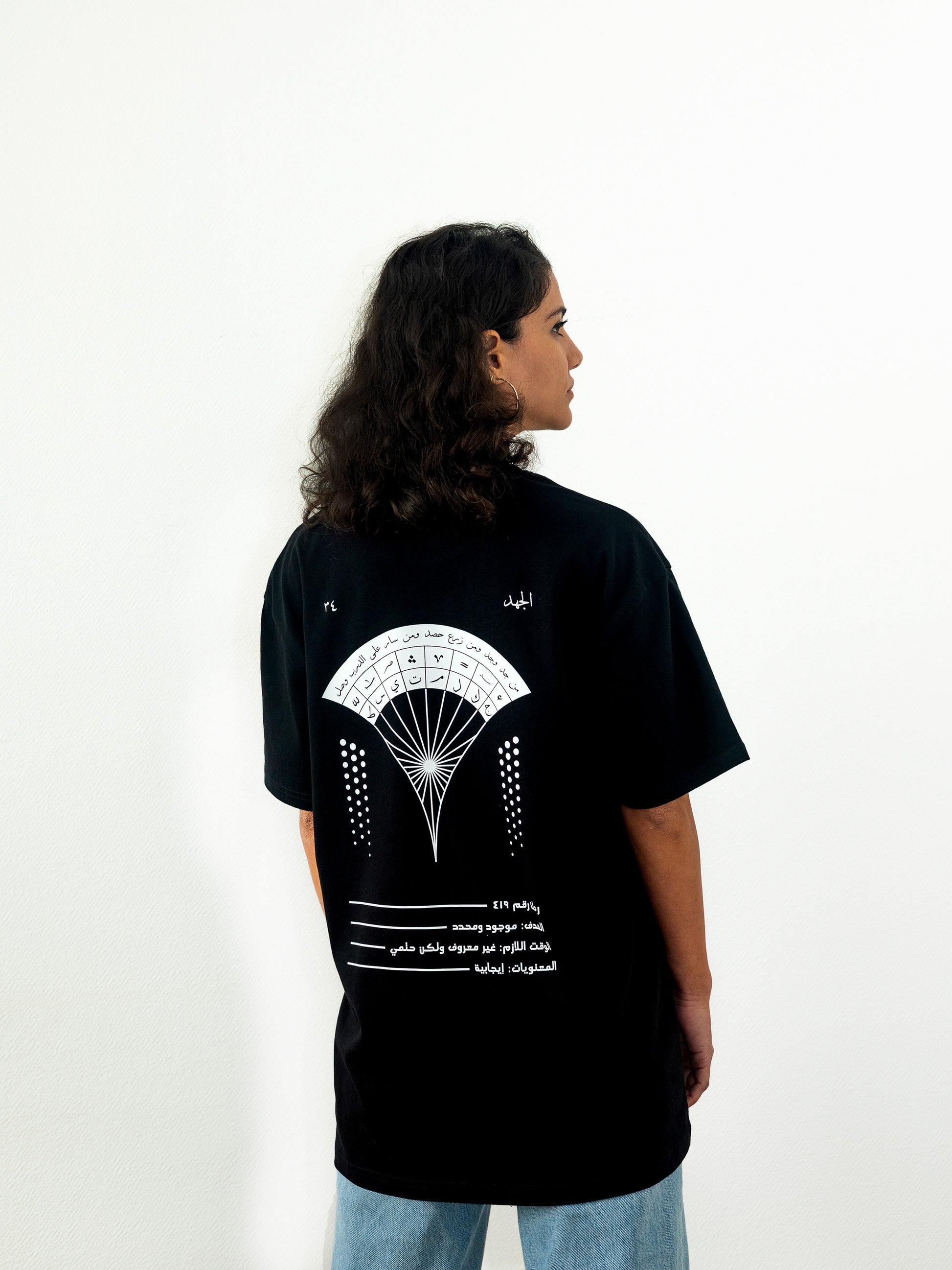 Backside view of beautiful arab model wearing a cool and creative over-sized t-shirt for streetwear and everyday lifestyle clothing. It features a beautiful arabic graphic print, emphasizing arabic art, culture and identity. It contains a famous arabic expression about hard work, perseverance and achievement. It uses arabic thuluth, a style of arabic calligraphy and lettering, including popular symbols. Black t-shirt. Backside view. Black and white print color.