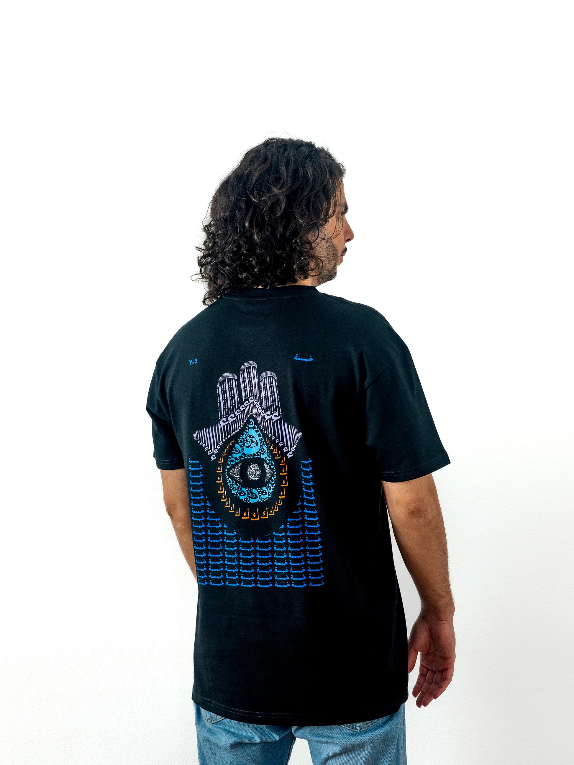 Beautiful arab male model wearing a cool and creative over-sized t-shirt for streetwear and everyday lifestyle clothing. This product features an exclusive print about khamsa, also known as hamsa, or the hand of Fatima. It is a popular amulet in Middle Eastern and North African culture, and is intended to protect against evil and envy. This art piece and pattern uses beautiful arabic letters and calligraphy to form its attractive shape. Black t-shirt. Backside view. Black and blue print color.