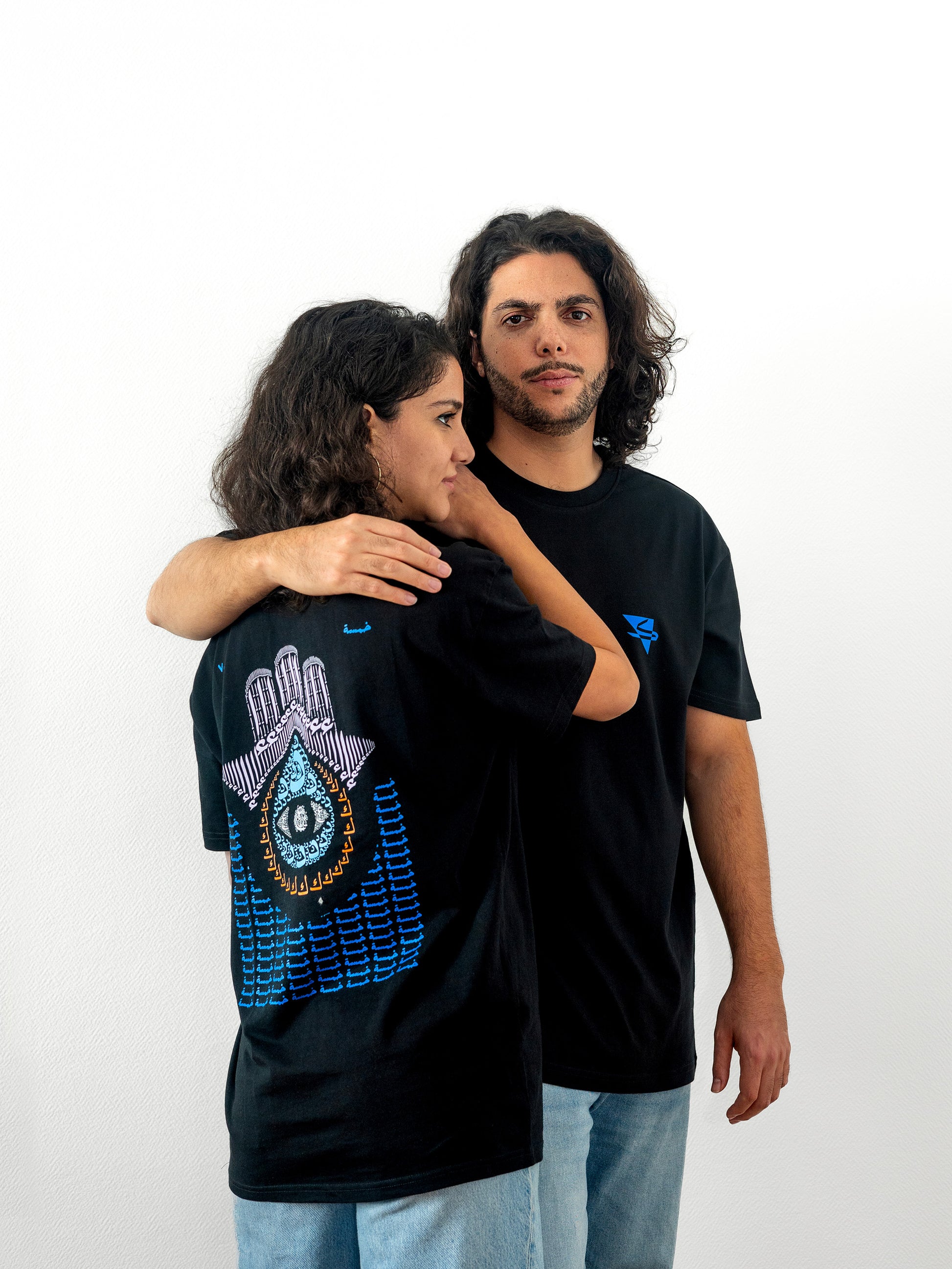 Two beautiful arab models wearing cool and creative over-sized t-shirts for streetwear and everyday lifestyle clothing. This product features an exclusive print about khamsa, also known as hamsa, or the hand of Fatima. It is a popular amulet in Middle Eastern and North African culture, and is intended to protect against evil and envy. This art piece and pattern uses beautiful arabic letters and calligraphy to form its attractive shape. Black t-shirt. Backside view. Black and blue print color.