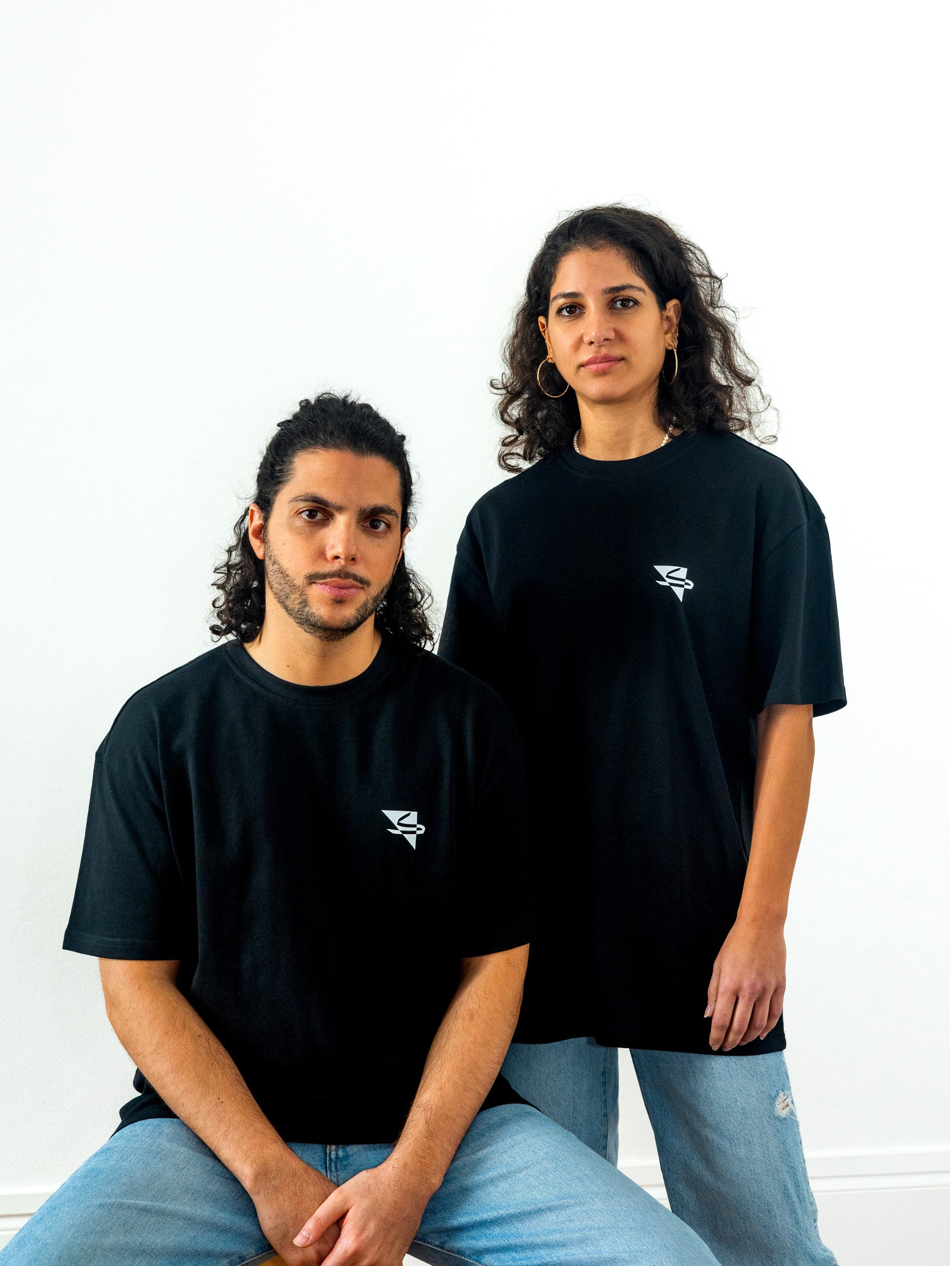 Two beautiful hot arab models wearing cool and creative over-sized t-shirts for streetwear and everyday lifestyle clothing. It features a simple beautiful logo, which is geometric in design and that has an arabic letter within it.  Black t-shirt. Frontside view. Black and white print color.