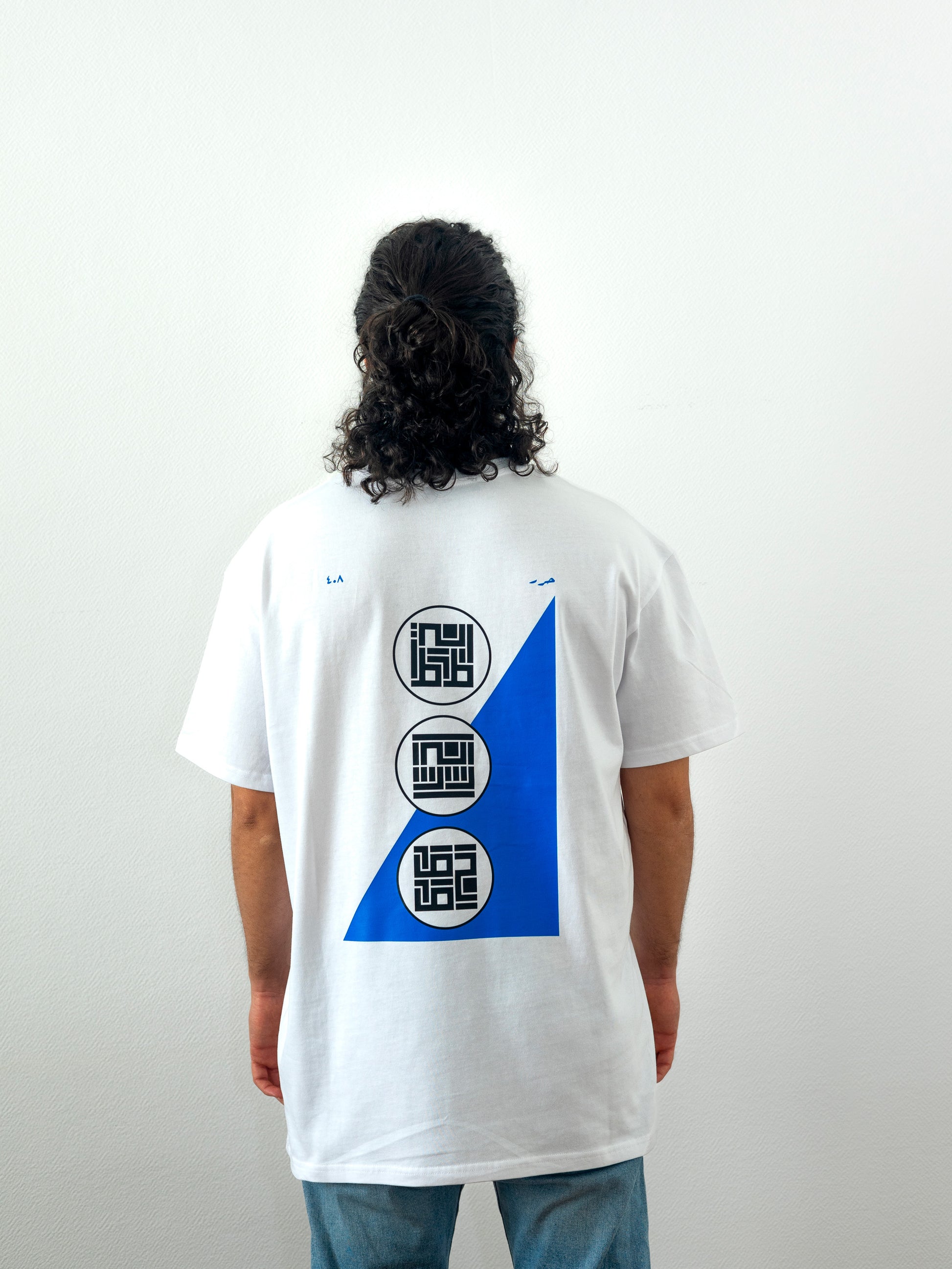 Backside of beautiful model wearing a cool and creative over-sized t-shirt for streetwear and everyday lifestyle clothing. It features an exclusive print emphasizing liberation of the individual and of society. Inspired by different types of arabic calligraphy, namely Kufic script, it exhibits a series of beautiful, geometrical circles and shapes. It is also draws from Japanese art and tribal design, highlighting the importance of spiritual freedom. White t-shirt. Backside view. Blue and black print color.