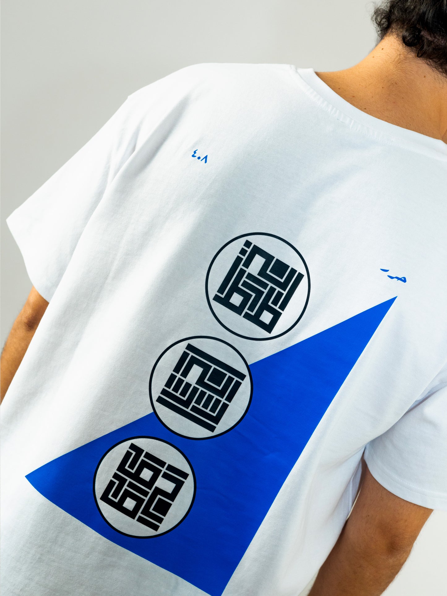 Close-up of a cool and creative over-sized t-shirt for streetwear and everyday lifestyle clothing. It features an exclusive print emphasizing liberation of the individual and of society. Inspired by different types of arabic calligraphy, namely Kufic script, it exhibits a series of beautiful, geometrical circles and shapes. It is also draws from Japanese art and tribal design, highlighting the importance of spiritual freedom. White t-shirt. Backside view. Blue and black print color.
