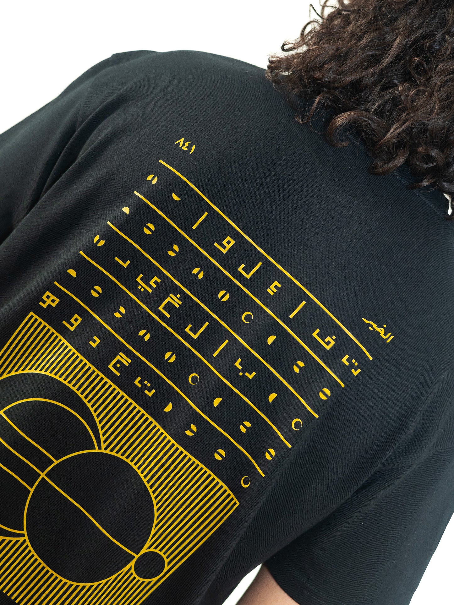 A cool and creative over-sized t-shirt for streetwear and everyday lifestyle clothing. This product features an exclusive print about a popular, common arabic expression regarding hope and optimism. Inspired by different types of arabic calligraphy, it exhibits a beautiful, geometrical style, featuring popular symbols such as the crescent, moon and stars. Black t-shirt. Backside view. Black, yellow, green and gold print color. Close-up.