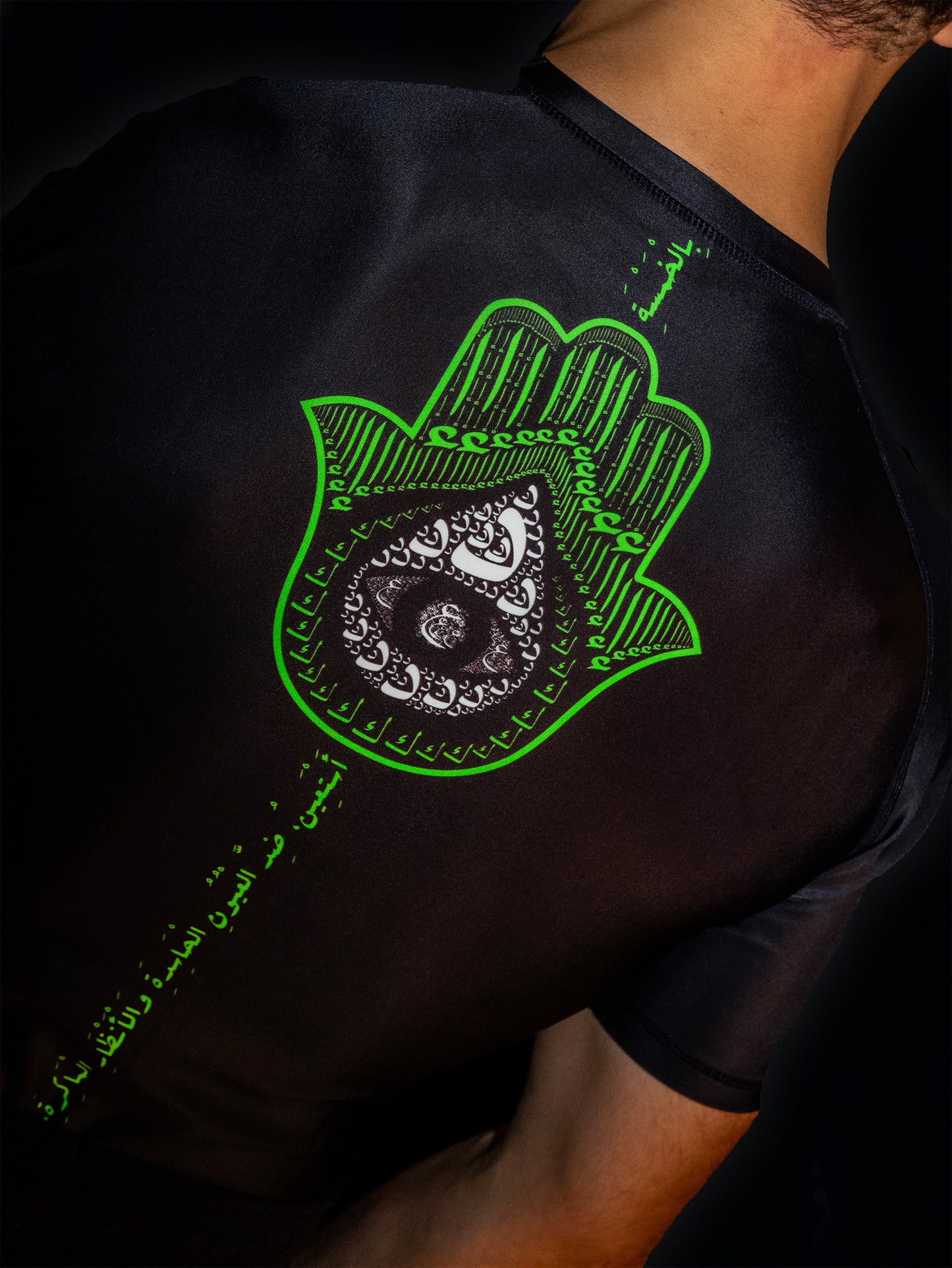 Black rashguard with arabic design, used for jiu-jitsu and other martial arts (e.g. MMA). This product features an exclusive print about khamsa, also known as hamsa, or the hand of Fatima. It is a popular amulet in Middle Eastern and North African culture, and is intended to protect against evil and envy. This art piece and pattern uses beautiful arabic letters and calligraphy to form its attractive shape. Backside view. Yellow and white print color. Arab model wearing it. Backside close-up