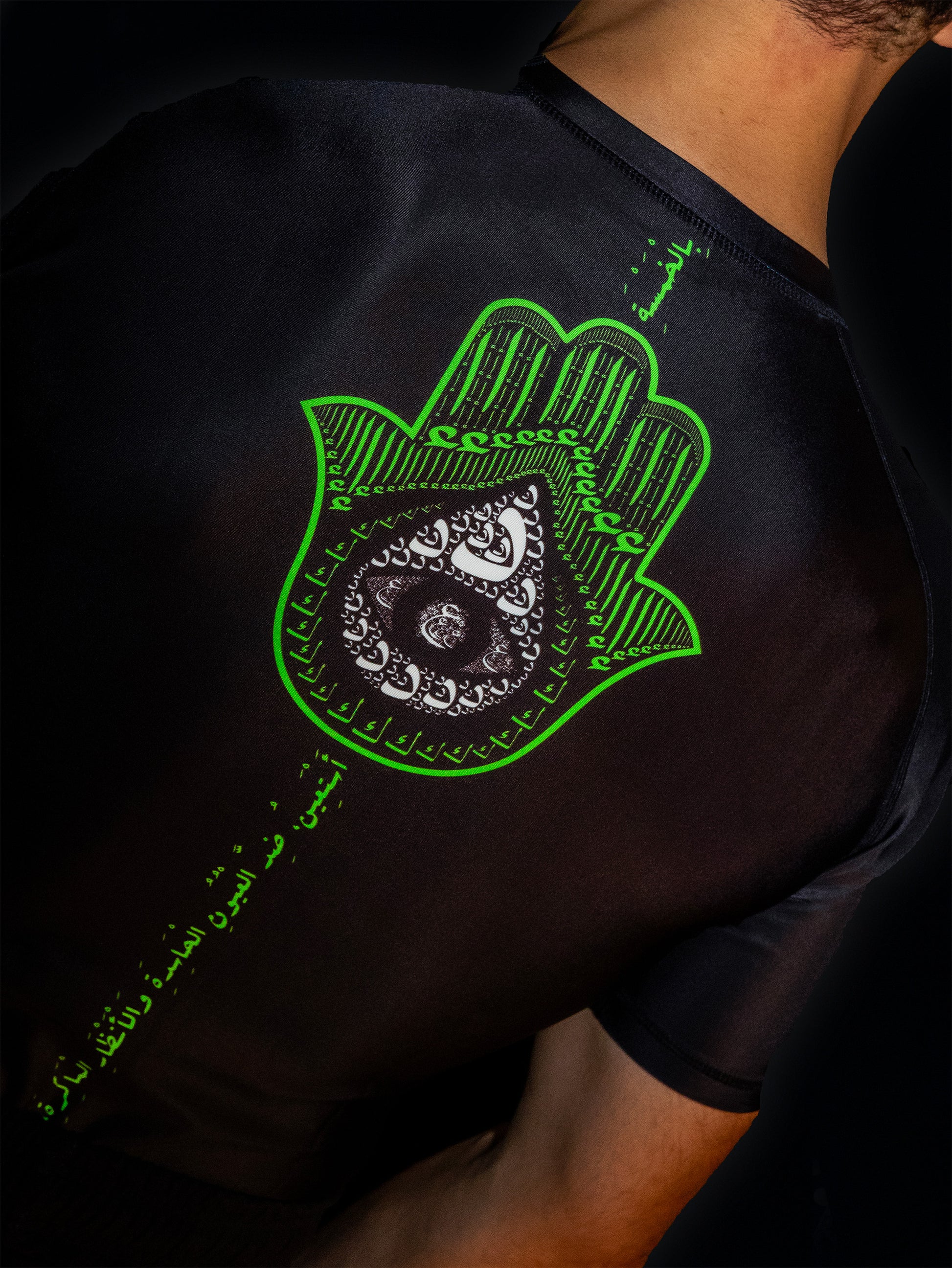 Black rashguard with arabic design, used for jiu-jitsu and other martial arts (e.g. MMA). This product features an exclusive print about khamsa, also known as hamsa, or the hand of Fatima. It is a popular amulet in Middle Eastern and North African culture, and is intended to protect against evil and envy. This art piece and pattern uses beautiful arabic letters and calligraphy to form its attractive shape. Backside view. Yellow and white print color. Arab model wearing it. Backside close-up