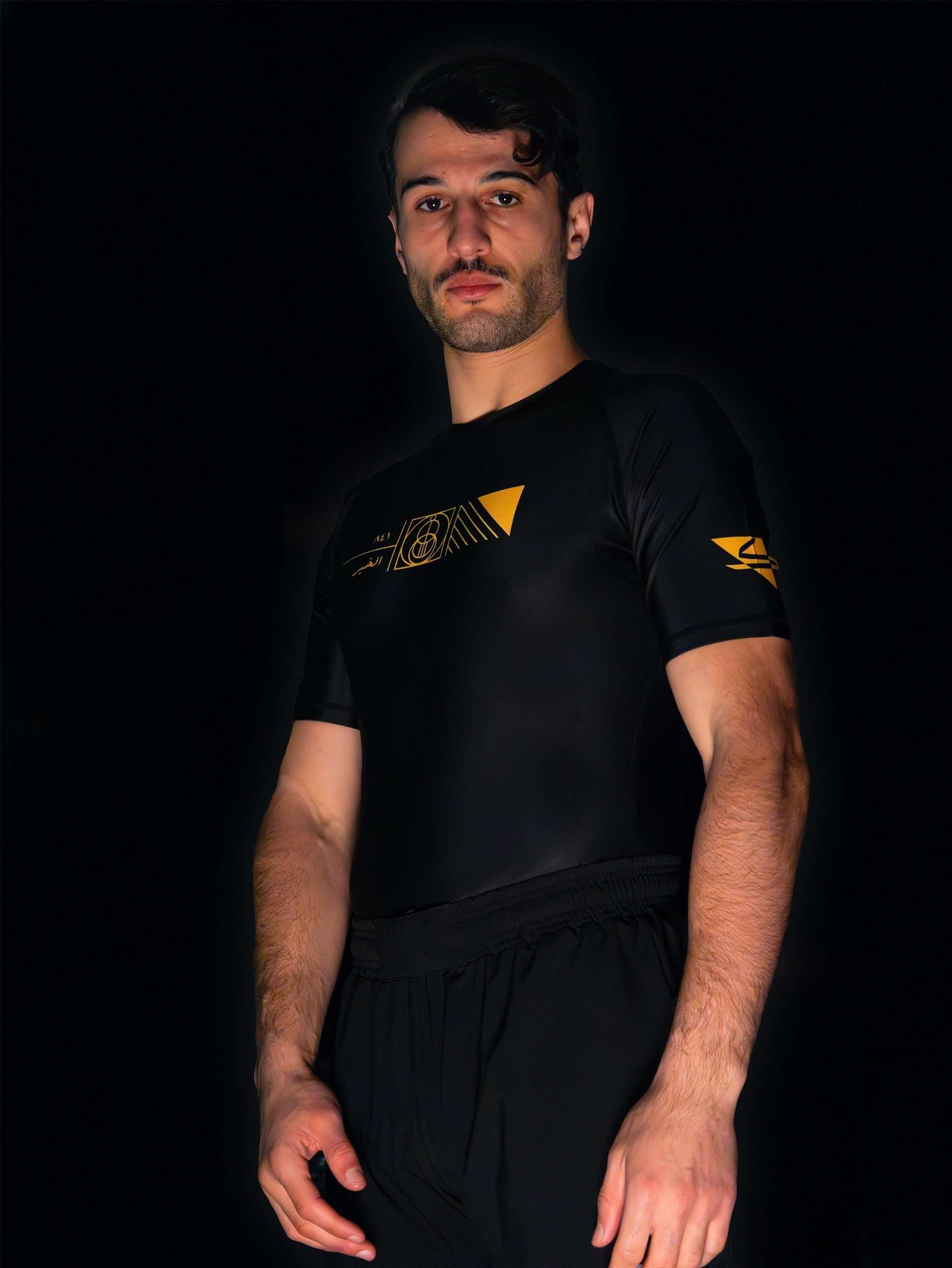 Black rashguard with arabic design, used for jiu-jitsu and other martial arts (e.g. MMA). This product features an exclusive print about a popular, common arabic expression regarding hope and optimism. Inspired by different types of arabic calligraphy, it exhibits a beautiful, geometrical style, featuring popular symbols such as the crescent, moon and stars. Frontside view. Yellow and Green print color. Left side angle.