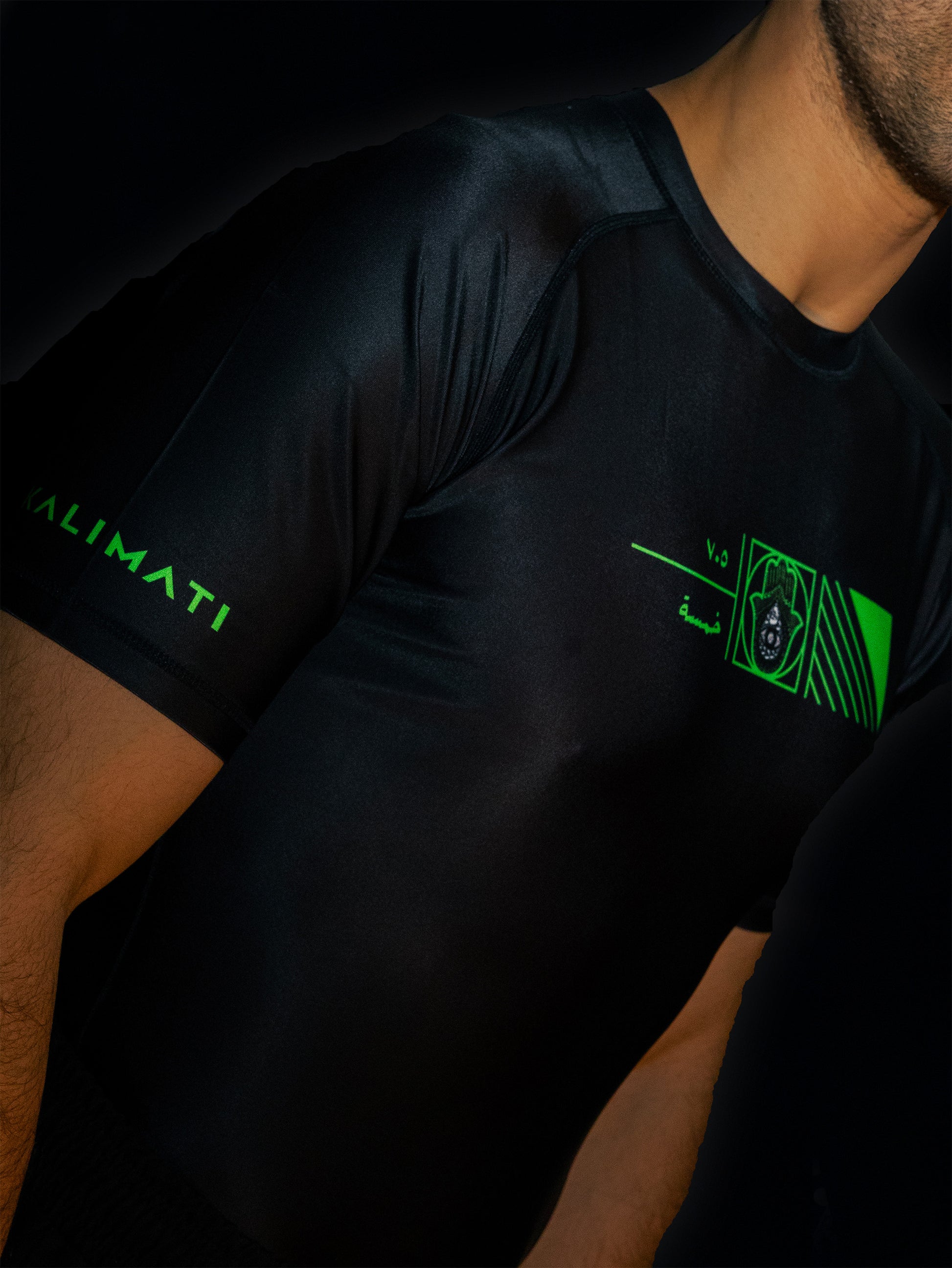 Black rashguard with arabic design, used for jiu-jitsu and other martial arts (e.g. MMA). This product features an exclusive print about khamsa, also known as hamsa, or the hand of Fatima. It is a popular amulet in Middle Eastern and North African culture, and is intended to protect against evil and envy. This art piece and pattern uses beautiful arabic letters and calligraphy to form its attractive shape. Yellow and white print color. Arab model wearing it. Sideaways close-up