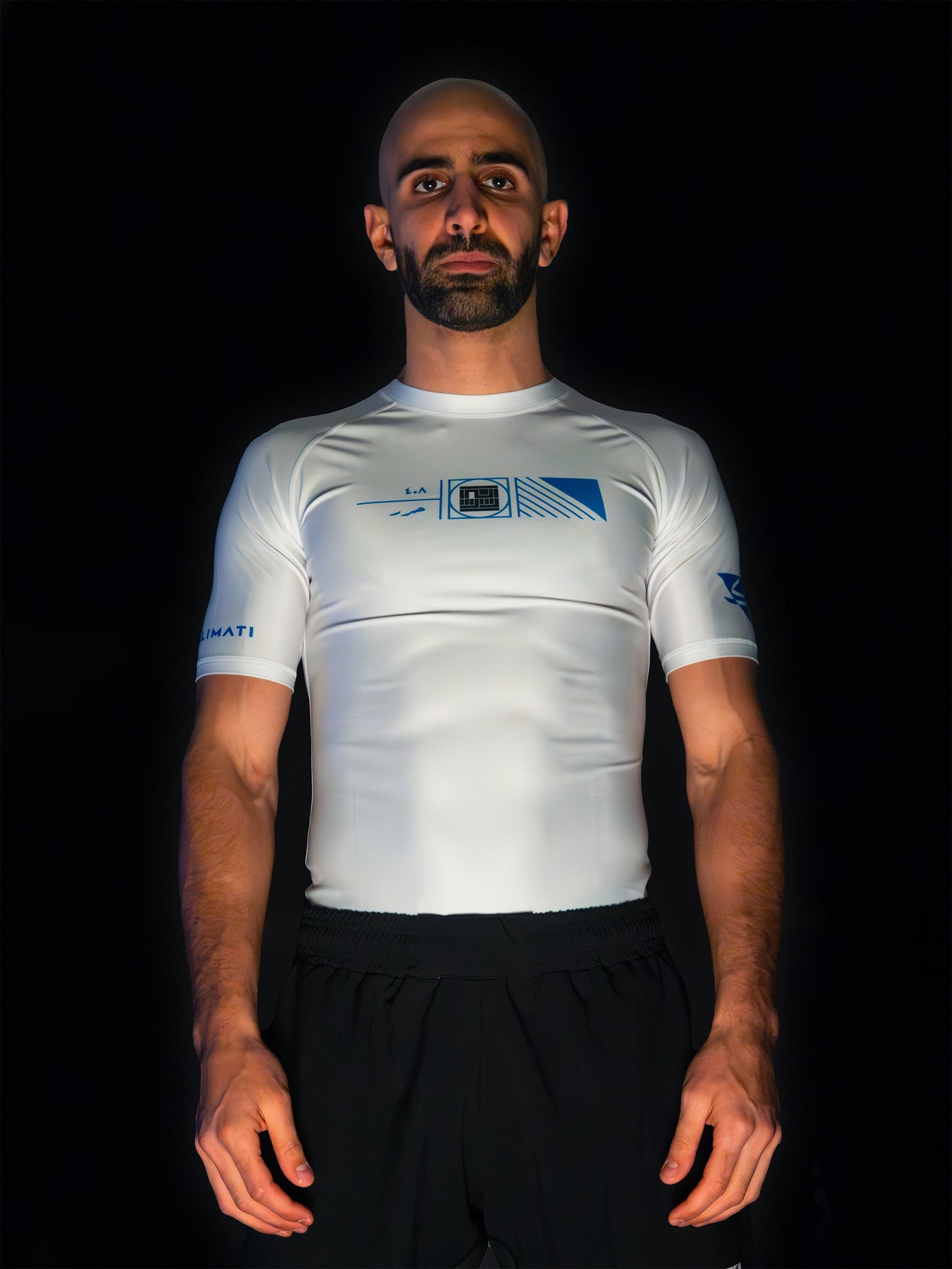 Arabic fighter wearing white rashguard with arabic print, used for jiu-jitsu and other martial arts (e.g. MMA). This product features an exclusive print emphasizing liberation of the individual and of society. Inspired by different types of arabic calligraphy, namely Kufic script, it exhibits a series of beautiful, geometrical circles and shapes. It is also draws from Japanese art and tribal design, highlighting the importance of spiritual freedom. Front view, standard photo. Model also wearing shorts.