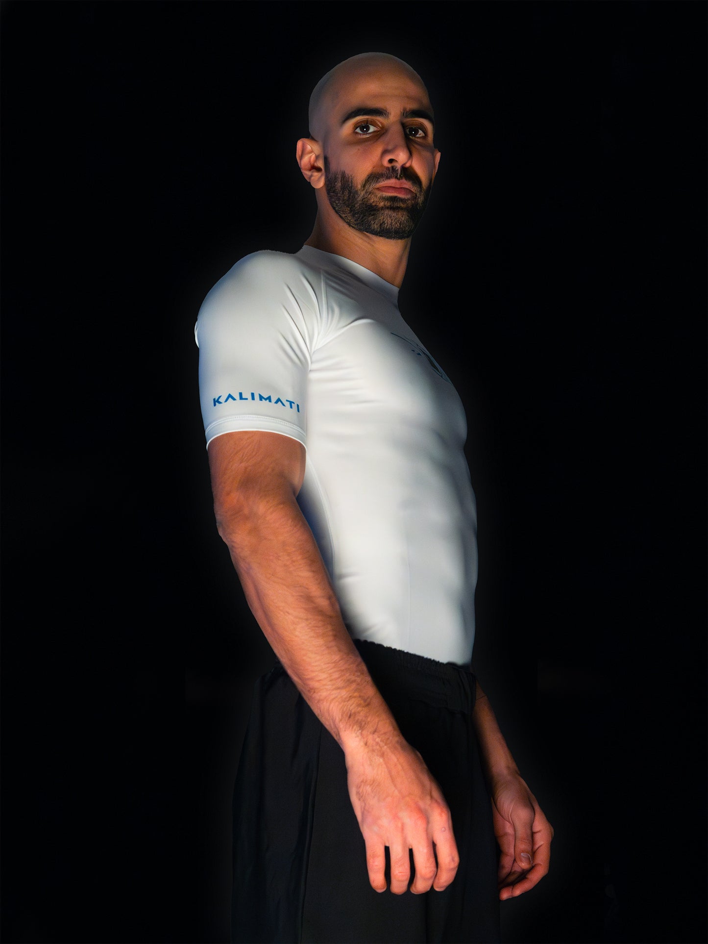 Arabic fighter wearing white rashguard with arabic print, used for jiu-jitsu and other martial arts (e.g. MMA). This product features an exclusive print emphasizing liberation of the individual and of society. Inspired by different types of arabic calligraphy, namely Kufic script, it exhibits a series of beautiful, geometrical circles and shapes. It is also draws from Japanese art and tribal design, highlighting the importance of spiritual freedom. Side view. Blue and black print color.